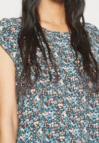Top à manches courtes à fleurs en bleu canard, marron et blanc. Il présente un col rond et un tissu léger et texturé avec un motif de fleurs éparpillées.