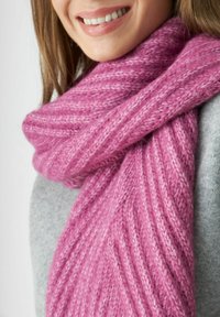 Glunderende persoon met een dikke, geribbelde roze gebreide sjaal om hun nek en een grijsgaren trui.