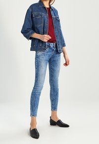 Chaqueta de denim con un corte relajado, pantalones vaqueros azules con un aspecto desgastado y zapatos negros planos. La parte superior es de color burdeos, añadiendo contraste al conjunto.