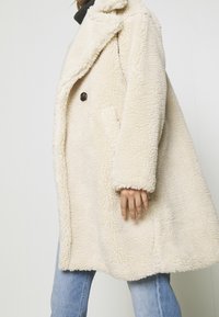 Personne portant un manteau crème à texture duveteuse avec un bouton noir, un jean bleu et un haut noir sur un fond clair uni.