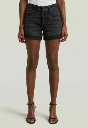 Vrouw die zwarte denim shorts met opgerolde zoom en zwarte sandaaltjes met enkelbandje draagt, staand tegen een effen lichte achtergrond.