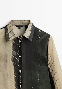 Chemise à manches longues boutonnée avec des motifs abstraits de coups de pinceau noirs et beiges et un col pointu sur fond blanc.