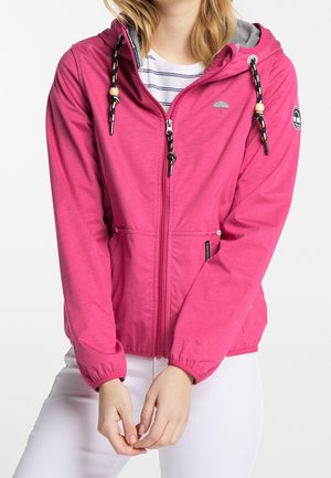 Femme portant une veste à capuche zippée rose vif avec des cordons noirs et jaunes par-dessus une chemise blanche rayée et un pantalon blanc.