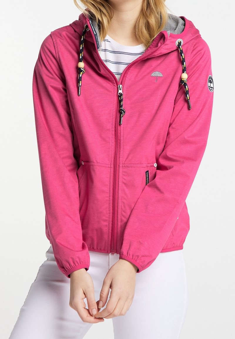 Femme portant une veste à capuche zippée rose vif avec des cordons noirs et jaunes par-dessus une chemise blanche rayée et un pantalon blanc.
