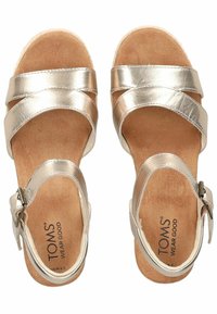 Metallic gold sandals with cross strap design, adjustable ankle buckle, cushioned footbed, and textured sole. Brand name "TOMS" visible.