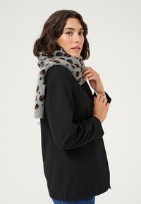 Blazer nero con una silhouette aderente, indossato con una sciarpa grigia con un motivo a leopardato nero e arancione. Tessuto liscio e texture sovrapposta.