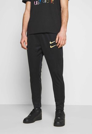 Mann trägt schwarze Nike-Sweatpants mit zwei goldenen Swoosh-Logos am linken Oberschenkel, schwarze Sneakers und ein schwarzes T-Shirt mit mehrfarbigem Text.