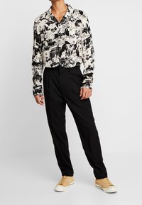 Gemustertes langärmliges Hemd in Schwarz und Weiß mit tropischem Design, kombiniert mit schwarzen Hosen und braunen Sneakers.