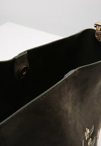 Sac à main en cuir vert foncé avec un motif floral brodé, texture lisse, fermeture à clip en métal, et doublure intérieure visible.