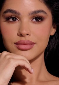 Primer plano de un rostro con piel suave, labios mauve plenos, cejas definidas y ojos marrones oscuros. El cabello es oscuro y ligeramente ondulado.