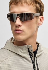 Sport sunglasses with a gray frame and dark lenses. Accompanied by a light gray jacket featuring a textured pattern and a black zipper.