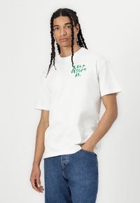 White cotton t-shirt with short sleeves featuring green graphic text on the chest; paired with blue denim jeans, standing against a white background.