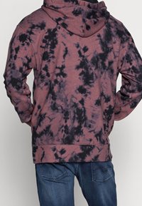 Sudadera color burdeos con patrón de tie-dye negro, que cuenta con un bolsillo canguro frontal y una capucha con cordón, fabricada con un tejido suave y texturizado.