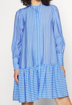 Niebieska sukienka z pionowymi i poziomymi białymi paskami, długimi bufkami na rękawach, zapinanymi na guziki mankietami oraz falbaną u dołu, noszona przez stojącą osobę.