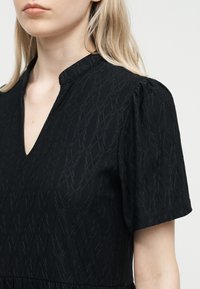 Γυναίκα με ξανθά μαλλιά, που φοράει μαύρο φόρεμα με υφή, κοντά μανίκια, λαιμόκοψη σε σχήμα V και ελαφρώς φουσκωτούς ώμους.