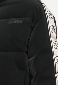 Sort puffjakke med mat finish, slankt design, sidelommer og en kontrasterende cremefarvet sideliste med sorte logo mønstre.
