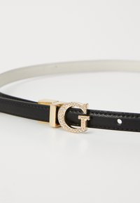 Zwarte leren riem met een gouden gesp waarop een gestileerde "G" staat en versierd met rhinestones, geplaatst tegen een eenvoudige witte achtergrond.