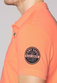 Polo de manga corta en color naranja brillante con una tela texturizada. Presenta un parche bordado redondo en la manga izquierda con texto circular.