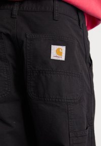 Pantalones cargo negros hechos de un tejido duradero, con múltiples bolsillos y un parche de logo cuadrado con un emblema amarillo en el bolsillo trasero.