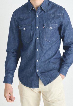 Chemise en denim foncé à manches longues avec col pointu, deux poches poitrine, boutons en argent et couture contrastante. Portée avec un pantalon de couleur claire.