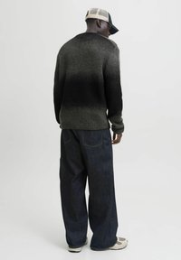 Ein Mann trägt einen schwarz-grau verlaufenden Pullover, lockere dunkelblaue Jeans, weiße Sneakers und eine schwarz-weiße Baseballkappe, und steht mit dem Rücken zu uns.