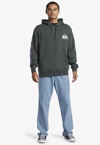 Felpa verde scuro con tasca frontale, cappuccio con cordino e logo blu sul petto. Abbinata a jeans azzurri chiari e sneakers nere.