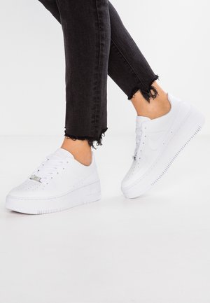 Personne portant des baskets blanches Nike Air Force 1 avec un jean noir à ourlet effiloché sur fond blanc.