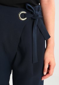 Granatowe spodnie z gładkiego materiału z szerokim pasem w talii i przednim wiązaniem, ozdobione dużym srebrnym pierścieniem D.