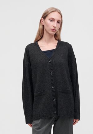 Donna con capelli biondi lunghi indossa un cardigan grigio scuro e morbido con bottoni sopra una maglia nera e pantaloni grigi, in piedi contro uno sfondo chiaro e semplice.