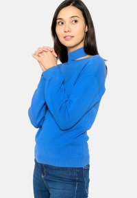 Pull en maille bleu avec un col montant et des découpes. Poignets et ourlet côtelés, fabriqué en tissu texturé doux. Porté avec un jean foncé.