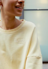 Žlutá mikina se žebrovaným límcem a vyšitým logem "Von Dutch Originals" na hrudi. Měkká látka s uvolněným střihem.