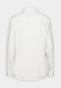 Chemise blanche à manches longues boutonnée vue de dos, avec un col classique et un design simple et sans ornement, sur fond uni.