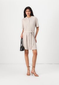 Knee-length beige dress with short sleeves, button closure, and fabric belt. Paired with strappy heels and a small black bag. Smooth texture.