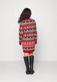 Pull en maille avec motifs festifs en rouge, noir, vert et blanc. Manches longues, ourlet arrondi, porté avec des bottes noires montantes jusqu'aux genoux.