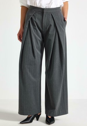 Donna che indossa pantaloni grigi a righe sottili a vita alta, con gamba larga e pieghe frontali, abbinati a scarpe nere con tacco basso e punta appuntita.
