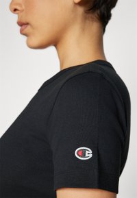 Sort bomulds T-shirt med korte ærmer. Har et lille, broderet logo på venstre ærme i blå, rød og hvid.