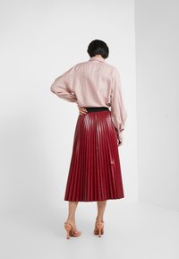 Rosa gestreiftes Hemd mit lockerer Passform, kombiniert mit einem glänzenden roten, plissierten Rock. Glatte Textur, schwarzer Taillenbund und Riemchenpumps.