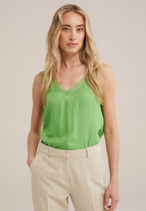 Grüne Satin-Camisole mit Spitzenbesatz entlang des V-Ausschnitts, kombiniert mit cremefarbenen Hosen. Glatte Textur, ärmelloses Design.
