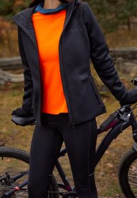 Atletisk outfit bestående av en svart jacka med dragkedja, en ljusorange tröja och svarta cykelbyxor med vadderad sittdyna, tillsammans med en cykel.