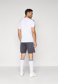 Bărbat în picioare, întors, purtând o cămașă sport albă, pantaloni scurți gri închis cu accente roșii, șosete albe până la genunchi și pantofi sport albi.