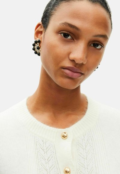 Hvid cardigan med ribbet krave, med guldknapper og en tekstureret chevron-mønster. Sorte statement-øreringe med sølvdetaljer.