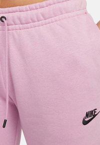 Ljusrosa sweatshirtbyxor i mjukt tyg med elastisk midja, snörning och en svart Nike-logotyp på vänster lår.