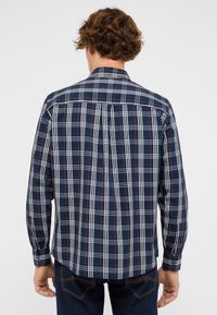 Mann mit lockigem Haar, der ein langärmliges, kariertes Hemd in Marineblau, Weiß und Rot sowie dunkle Jeans trägt, von hinten vor einem schlichten Hintergrund gezeigt.