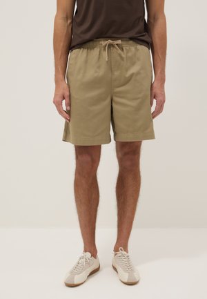 Mann trägt beige Shorts mit Kordelzug, ein dunkelbraunes Hemd und weiße Turnschuhe und steht vor hellem Hintergrund.