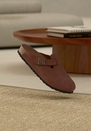Zapato tipo slip-on de ante marrón con una hebilla, flotando sobre una alfombra beige texturizada, junto a una mesa de café redonda de madera con libros apilados.