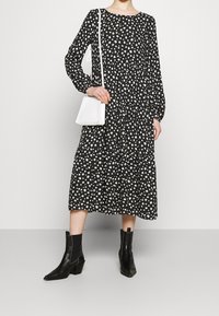 Musta pitkähihainen mekko, jossa on valkoinen kukkakuvio, kerroksellinen muotoilu, yhdistettynä valkoiseen olkibag- laukkuun ja mustiin nilkkureihin, joissa on paksut korot.