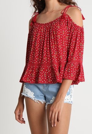 Blusa roja con los hombros descubiertos, estampado floral, dobladillo con volantes y tiras para atar. Combinada con shorts de mezclilla azul claro con detalle de encaje.