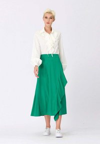 Blouse blanche avec un devant à volants et des manches bouffantes, associée à une jupe plissée verte à mi-mollet, avec un design en couches et des baskets blanches.