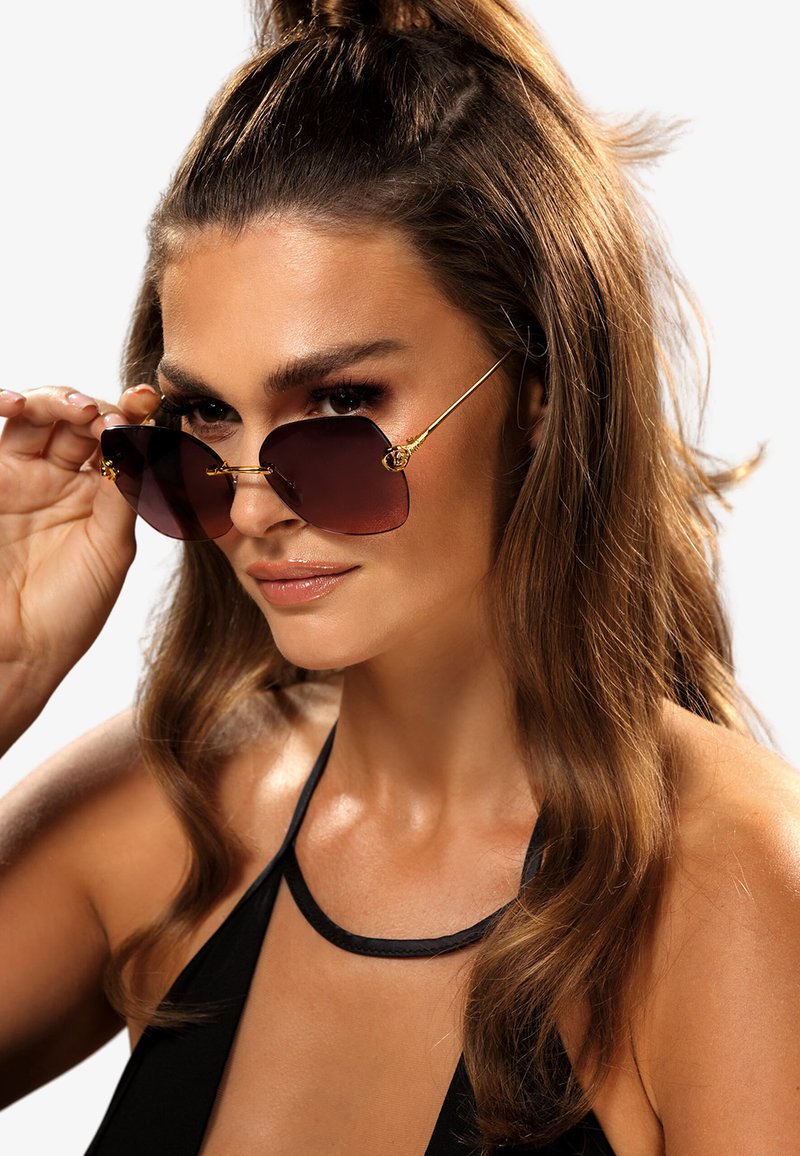 Okulary przeciwsłoneczne z ciemno-brązowymi, kanciastymi szkłami i złotymi metalowymi akcentami. Ramka ma unikalny design z dekoracyjnymi elementami.