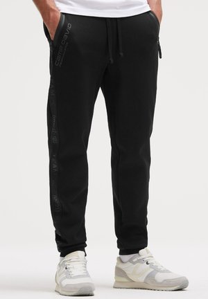 Mann trägt schwarze Cam David Jogginghosen mit Text an der Seite und weiß-graue Sneakers, Hände in den Taschen, steht vor schlichtem Hintergrund.